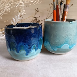 Op de afbeelding: Twee keramische bekers met een ronde vorm en een inkeping in de rand. De linkerbeker heeft een diepblauw glazuur dat uitloopt in een lichtere turquoise basis. De rechterbeker heeft een lichtgroen craquelé glazuur met een turquoise druipeffect.