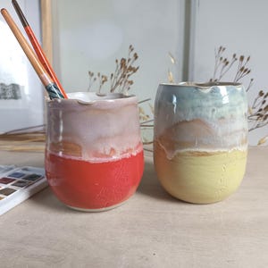Può includere: Due bicchieri in ceramica dalla forma arrotondata e finitura lucida. Il bicchiere a sinistra presenta una base rosso vivace con uno smalto colato rosa e bianco. Il bicchiere a destra ha una base giallo pallido con uno smalto colato blu e bianco.
