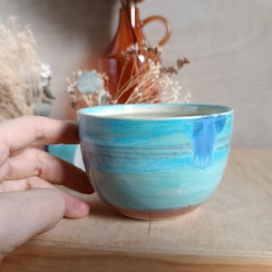 May include: A handmade ceramic bowl with a turquoise and blue glaze. The bowl has a rounded shape and a brown base. The bowl is held by a hand and is sitting on a wooden surface. Decorative items are in the background.