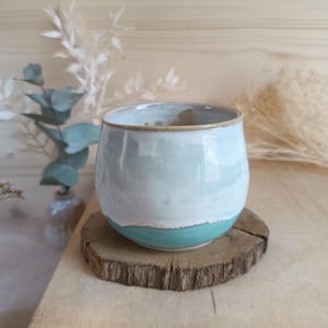 Peut inclure: Une tasse en céramique faite à la main avec un glaçage bleu clair et blanc, posée sur un dessous de verre en bois rustique. La tasse a une forme arrondie et une base turquoise. Le bord est orné d'une bande dorée. Des plantes décoratives sont en arrière-plan.