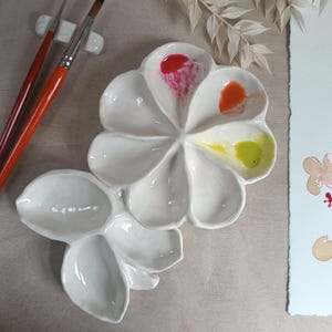 Pode incluir: Uma paleta de pintura branca em cerâmica em forma de flor com tinta vermelha, laranja e amarela. Dois pincéis com cabos laranja repousam sobre um pequeno suporte branco. Uma aquarela com folhas verdes e flores cor-de-rosa está à direita.