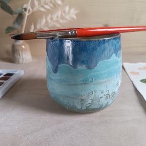 Puede incluir: Un vaso de cerámica con un esmalte azul y turquesa, que sostiene un pincel con mango rojo. Una paleta de acuarelas y un diseño floral pintado están cerca. El vaso tiene una superficie texturizada y un efecto de esmalte goteado.