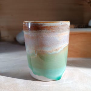 Peut inclure: Une tasse en céramique cylindrique avec un émail aux tons multiples. Le bord supérieur est brun foncé, passant par une section centrale lavande et crème, pour finir sur une base vert vif qui coule vers le bord inférieur de la tasse.