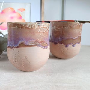 Può includere: Due tazze in ceramica con una base opaca color pesca chiaro e una finitura smaltata lucida e colata nei toni del marrone, viola e bianco. Le tazze presentano una superficie ruvida e un piccolo beccuccio sul bordo. Un pennello è appoggiato sopra.