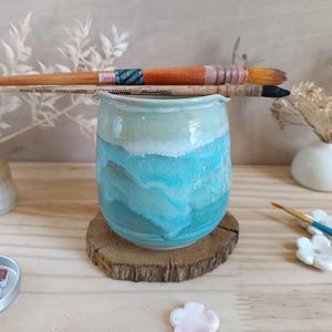Könnte beinhalten: Ein handgefertigter Keramik-Kunstbedarfshalter in Blau- und Grüntönen, der auf einer Holzscheibe steht. Zwei Pinsel liegen obenauf. Weiteres Kunstzubehör und dekorative Elemente befinden sich im Hintergrund.