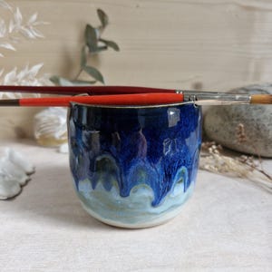 Peut inclure: Une tasse en céramique avec un vernis brillant présentant un sommet bleu profond qui se transforme en un motif de gouttes bleu clair et blanc. Deux pinceaux à manche rouge reposent horizontalement sur le bord supérieur de la tasse.