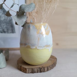 Op de afbeelding: Een keramieken vaas met een ronde, eivormige vorm, voorzien van een matte gele basis en een glanzende, druipende witte en goudgele glazuurlaag aan de bovenzijde. De vaas bevat gedroogde eucalyptusstengels en dunne, lichtgekleurde siergrassen.