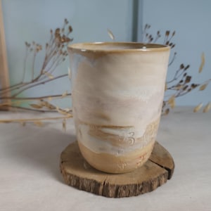 Op de afbeelding: Een cilindervormige keramische beker met een gladde, gebroken witte en beige geglazuurde afwerking met subtiele, gelaagde kleurvariaties. Het onderste gedeelte van de beker vertoont vage, in reliëf aangebrachte decoratieve markeringen.