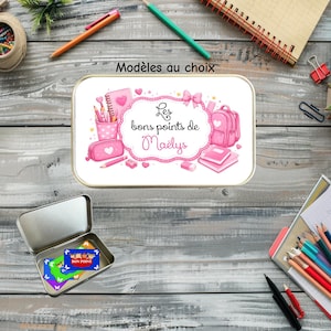 Peut inclure: Une boîte en métal rectangulaire avec un motif graphique rose représentant des fournitures scolaires et le texte "Les bons points de Maëlys". Une boîte en métal ouverte se trouve en dessous, contenant des petites cartes rectangulaires colorées à motifs de papillons.