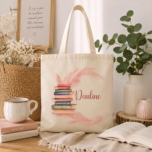 Peut inclure: Un sac fourre-tout en toile couleur crème avec une illustration imprimée d'une pile de livres colorés entourée de plumes roses douces. Le prénom Pauline est imprimé en écriture cursive à côté des livres. Le sac possède deux anses solides.