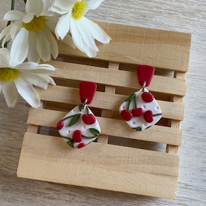 May include: A pair of red and white cherry-themed earrings with a white background and green leaves. The earrings are shaped like a diamond and have a red clay base.
