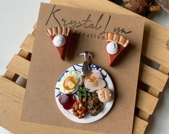 May include: A set of Thanksgiving-themed polymer clay earrings and a necklace. The earrings are shaped like pumpkin pies with a white dollop of whipped cream on top. The necklace features a plate with a miniature Thanksgiving dinner, including mashed potatoes, gravy, stuffing, turkey, green beans, cranberry sauce, and a dinner roll.