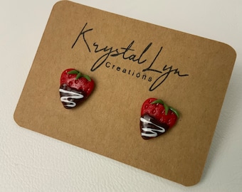 Può includere: Un paio di orecchini a forma di fragola ricoperti di cioccolato rosso e marrone. Gli orecchini sono realizzati in pasta polimerica e hanno una glassa bianca in cima.