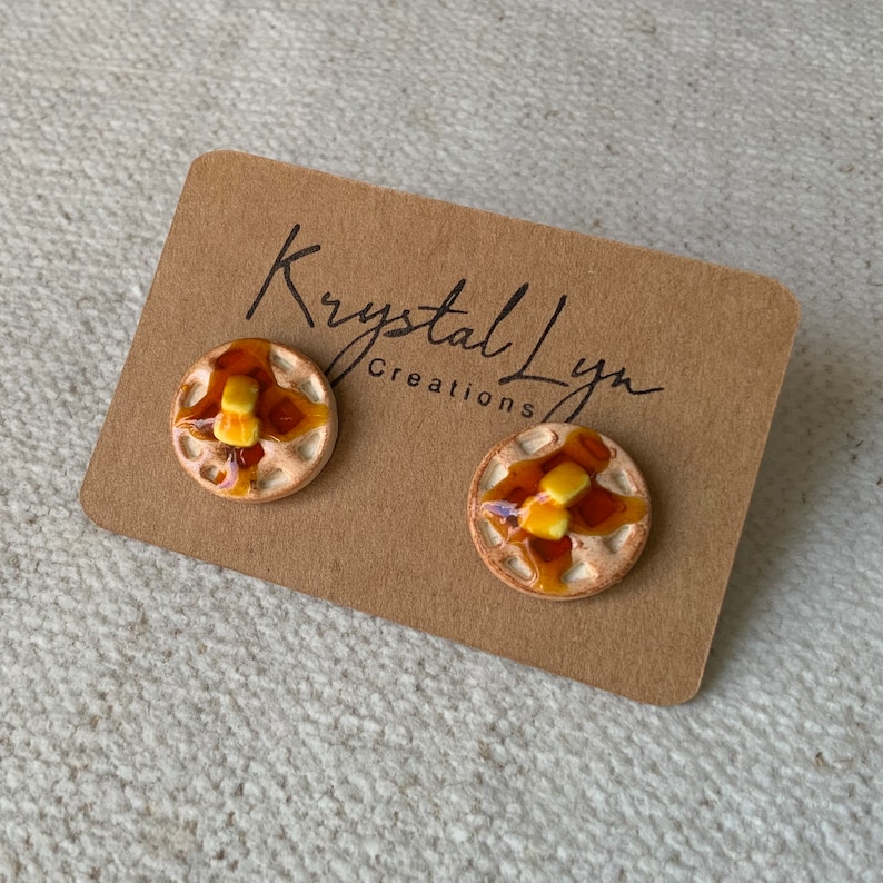 May include: A pair of ceramic waffle earrings with a brown glaze and a yellow glaze for the syrup and butter. The earrings are on a brown card with the text "Krystal Lyn Creations".