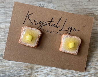 Puede incluir: Un par de pendientes hechos a mano con forma de rebanadas de tostadas con una porción de mantequilla en la parte superior. Los pendientes están en una tarjeta marrón con el texto "Krystal Lyn Creations".