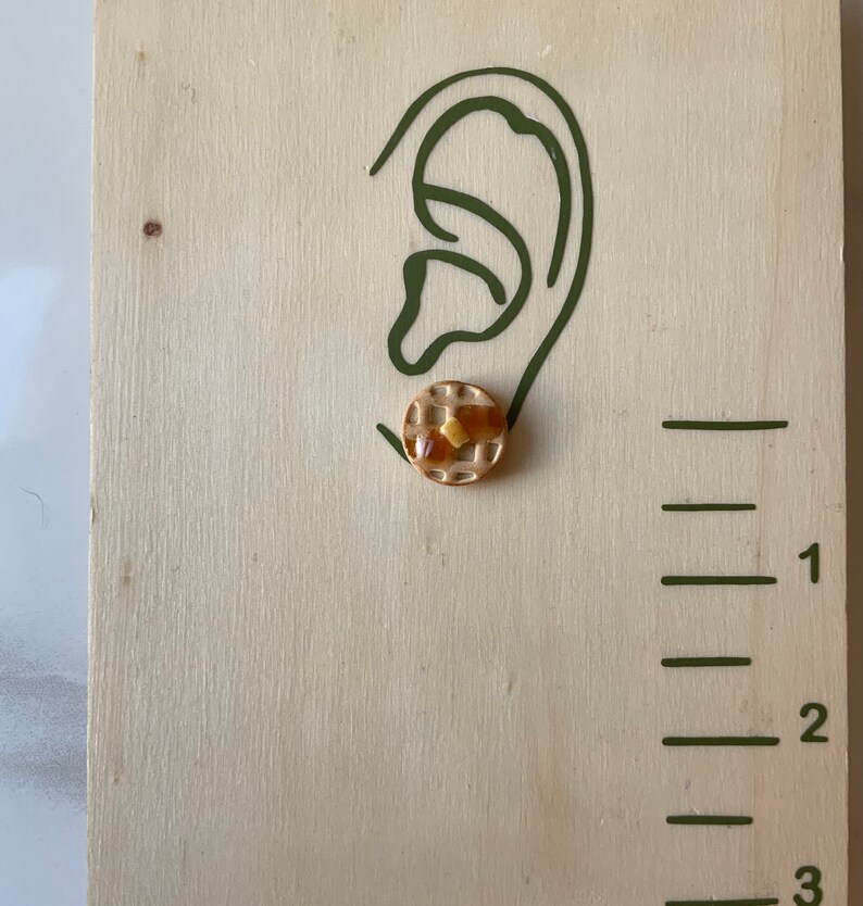 May include: A pair of gold-tone stud earrings shaped like a waffle with a yellow syrup topping.