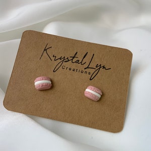 May include: A pair of pink and white macaron stud earrings. The earrings are on a brown card with the text "Krystal Lyn Creations".