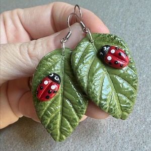 May include: A pair of handmade earrings featuring green leaf shapes with a glossy finish. Each leaf has a red ladybug with black spots and white accents. The earrings have silver-colored hooks.
