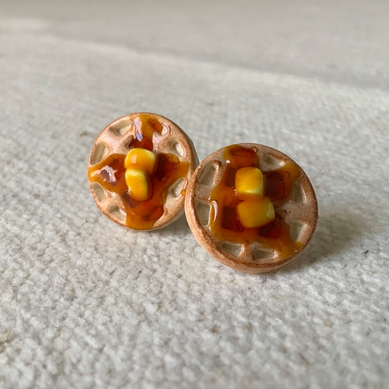May include: A pair of ceramic waffle earrings with a brown glaze and yellow butter topping. The earrings are small and round, and they are perfect for adding a touch of whimsy to any outfit.