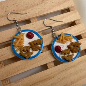 May include: A pair of novelty earrings featuring miniature food designs. Each earring showcases a white plate with a blue rim, adorned with tiny fries, chicken nuggets, and a dollop of red sauce. The earrings hang from silver-colored hooks.