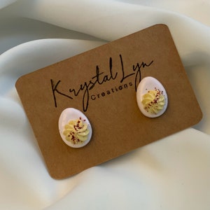 May include: A pair of white egg-shaped earrings with yellow and red decorative details. The earrings are on a brown card with the text "Krystal Lyn Creations".