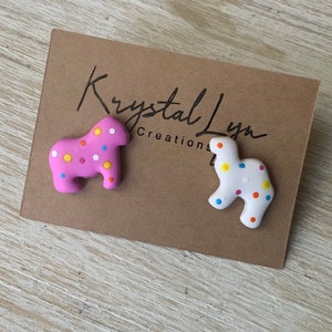 May include: Two colorful cookie-shaped earrings, one pink with white and multi-colored dots, and one white with multi-colored dots. The earrings are on a brown card with the text "Krystal Lynn Creations".