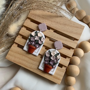 May include: A pair of handmade earrings featuring a floral design. Each earring has a white arch shape with a small terracotta-colored pot filled with flowers in shades of purple and cream. The earrings have a textured, square stud in a muted purple color.