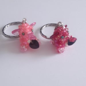 Peut inclure: Deux porte-clés représentant de petites figurines d'animaux en trois dimensions, fabriquées à partir de perles roses et magenta translucides. Chaque figurine possède une perle sombre en forme de cœur et un anneau en métal argenté.