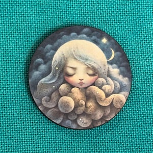 May include: A circular button with an illustration of a peaceful figure with closed eyes, encircled by cloud-like forms. The figure has long, flowing hair and is set against a dark, starry sky with a crescent moon. The button is on a teal fabric background.