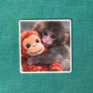 May include: A square sticker featuring a photographic print of a small, brown, furry monkey embracing a bright orange plush monkey toy. The sticker has a thin white border and is set against a solid, textured, dark teal fabric background.