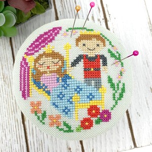 May include: A round, light green pincushion with a cross-stitched design of a princess and prince. The princess is in a blue dress, and the prince is wearing a red vest. The pincushion has three pink pins and is surrounded by a floral border.