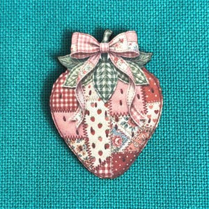 May include: A strawberry-shaped patch featuring a patchwork design of red, pink, and white fabric patterns including gingham, floral, and small strawberry prints. The strawberry is topped with green fabric leaves and a pink and white gingham bow.