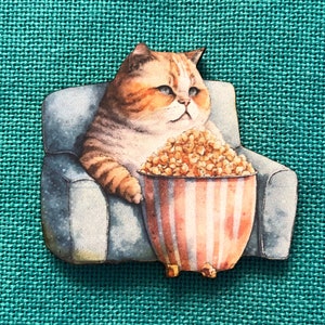 May include: A cat illustration sitting in a gray armchair, holding a striped popcorn bucket. The cat is tan and white with blue eyes. The popcorn bucket is red and white striped.