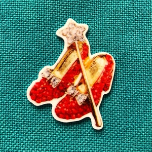 May include: A decorative pin featuring a pair of red shoes with a star-shaped wand. The shoes are embellished with glitter and small bows. The wand is gold with a star-shaped top.