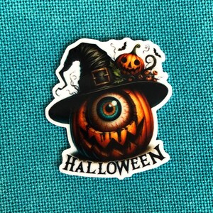 Puede incluir: Una calabaza de Halloween con un sombrero de bruja y un solo ojo. La calabaza es naranja y marrón con una cara de miedo. El sombrero es negro con un borde verde. El ojo es azul con un iris marrón. El texto "HALLOWEEN" está escrito debajo de la calabaza.
