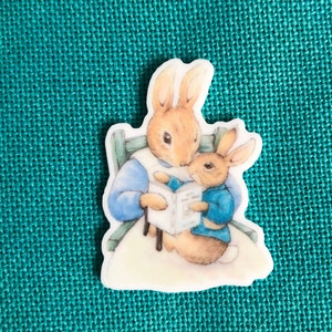 May include: A white ceramic button featuring a vintage illustration of two rabbits reading a book together. The mother rabbit is wearing a blue dress and the baby rabbit is wearing a blue shirt.