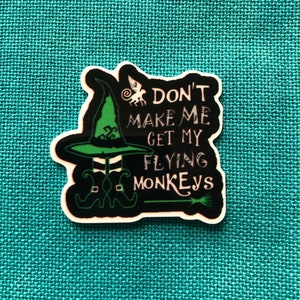 May include: A black and white sticker with the text "DON'T MAKE ME GET MY FLYING MONKEYS." The sticker features a green witch hat, witch legs, and a broom. The background is a teal fabric.
