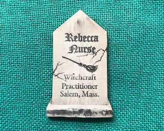 Könnte beinhalten: Graue, grabsteinförmige Anstecknadel mit dem Namen "Rebecca Nurse" in gotischer Schrift. Darunter eine Besen-Grafik und die Worte "Witchcraft Practitioner Salem, Mass." sind zu sehen. Die Anstecknadel befindet sich vor einem türkisfarbenen Stoffhintergrund.