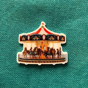 May include: A white ceramic carousel ornament with a red, white, and blue roof and gold accents. The carousel has horses and riders in various colors.