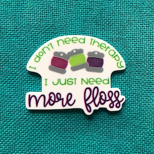 May include: A white sticker with the text "I don't need therapy, I just need more floss." The sticker features three spools of thread in pink, green, and purple. The background is a teal fabric.
