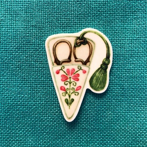 May include: A white ceramic scissor holder with a floral design and a green tassel. The holder is shaped like a cone and has a pair of scissors inside.