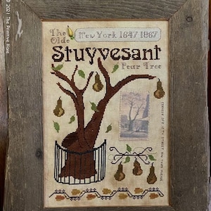 Op de afbeelding: Een ingelijst kruissteekborduurwerk met de tekst "The Olde Stuyvesant Pear Tree" en een afbeelding van een perenboom. Daaronder vier peren op een kanten doek. Het borduurwerk zit in een rustieke houten lijst.