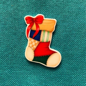 Puede incluir: Adorno en forma de calcetín navideño colorido con un lazo rojo. El calcetín presenta patrones de patchwork en rojo, verde, azul y amarillo. El fondo es una tela turquesa texturizada.