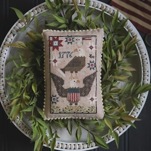 May include: A beige, rectangular, cross-stitched pillow featuring an eagle with outstretched wings, the text "1776", and patriotic stars and flags. The pillow is surrounded by a white beaded tray and green foliage.