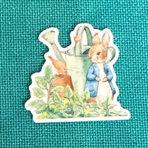 May include: A white sticker with a cartoon illustration of a rabbit holding a watering can. A small bird is perched on the watering can. The background is green with leaves and flowers.