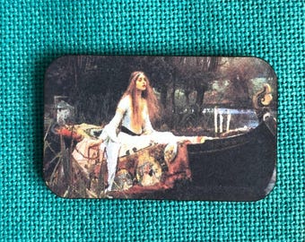 May include: A rectangular magnet featuring a reproduction of John William Waterhouse's painting The Lady of Shalott, depicting a woman with long hair in a white dress sitting in a dark boat on a river surrounded by trees and foliage.