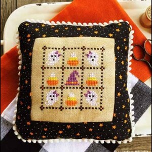 Puede incluir: Un cojín cuadrado decorativo con temática de Halloween. Presenta un diseño de punto de cruz de fantasmas, caramelos de maíz y un sombrero de bruja. El borde es negro con estrellas naranjas y ribete de pompón blanco. El texto en la parte superior dice "OCTOBER NINE PATCH".