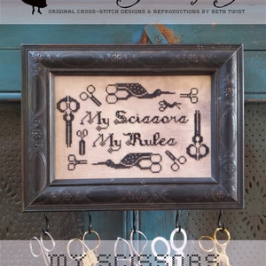 May include: A framed cross-stitch sampler with black thread on white fabric. The sampler reads "My Scissors My Rules" and features a design of scissors and birds. The frame is black with ornate detailing and is hanging on a blue wall with hooks and scissors hanging below.