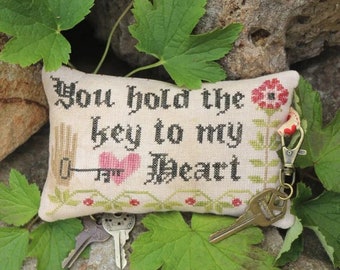May include: A cross-stitch sampler with the text "You hold the key to my heart" and a small heart and key design. The sampler is attached to a key ring with a key.