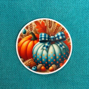 Puede incluir: Una pegatina redonda con temática de cosecha otoñal. Representa una calabaza naranja y una calabaza azul con lunares naranjas, un lazo a cuadros azul y blanco, bellotas y espigas de trigo sobre un fondo marrón.